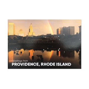 Könnte beinhalten: Eine Postkarte mit Blick auf die Skyline von Providence, Rhode Island bei Sonnenuntergang. Die Skyline der Stadt spiegelt sich im Wasser. Ein Regenbogen erscheint am Himmel über der Stadt.
