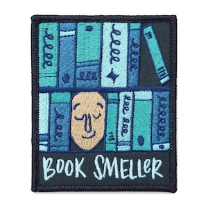 Op de afbeelding: Geborduurde strijk-op patch met een cartoon gezicht omringd door een stapel boeken. De tekst "BOOK SMELLER" staat onder de afbeelding.