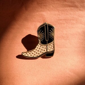 May include: A gold-toned metal cowboy boot pin with a black top and heel. The boot features a cream-colored design on the front and a sunburst pattern on the black top. The pin is set against a peach-colored background.
