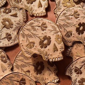 Puede incluir: Una colección de parches bordados con un diseño de calavera con detalles florales. Los parches son beige con bordados dorados y tienen una estética vintage.