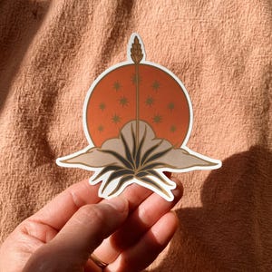 May include: A sticker with a stylized illustration of a plant with a tall stalk and a sun with a star pattern behind it. The plant is brown and black, the sun is orange, and the stars are white.