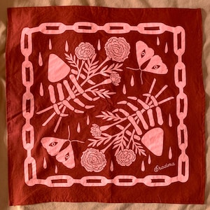 May include: A rust-colored bandana with a chain border and a repeating pattern of skeleton hands, flowers, moths, and raindrops in a lighter shade of red. The word "Eradura" is printed in the bottom right corner.