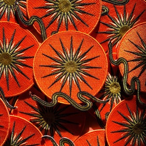 May include: Embroidered patches with a sunburst design. The patches have an orange background, a central gold circle, and black and white rays. A green and black embroidered snake winds around some of the patches, adding detail.