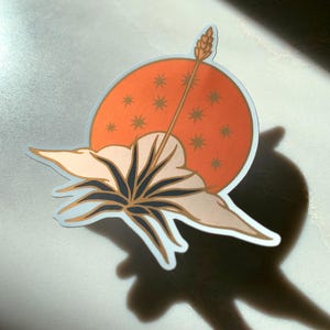 May include: A sticker with a stylized illustration of a flower with a large, white blossom and a long, thin stem. The flower is set against a background of a red circle with gold stars.