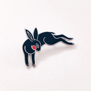 May include: Black enamel pin of a rabbit with red eyes, jumping in mid-air. The pin is silver-toned.