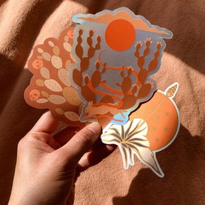 May include: Three illustrated stickers featuring desert scenes. The first sticker shows a red sun setting over a blue sky with cacti in the foreground. The second sticker shows a brown desert landscape with cacti and a red ribbon. The third sticker shows a white flower with a brown stem and a red sun with stars in the background.