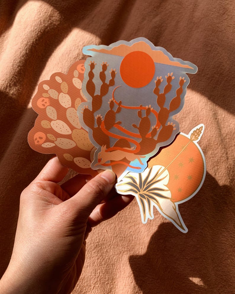 May include: Three illustrated stickers featuring desert scenes. The first sticker shows a red sun setting over a blue sky with cacti in the foreground. The second sticker shows a brown desert landscape with cacti and a red ribbon. The third sticker shows a white flower with a brown stem and a red sun with stars in the background.