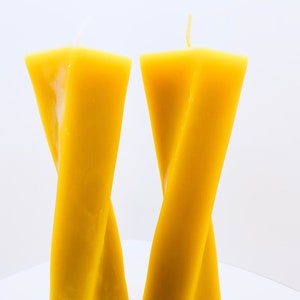 May include: Two yellow beeswax candles twisted into a unique shape. The candles are standing upright on a white surface. A gold ring is in the foreground.