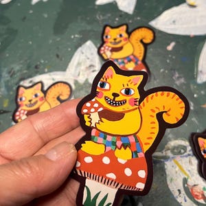 May include: A collection of whimsical stickers featuring a yellow cat-squirrel hybrid character. The character is holding a mushroom and wearing a colorful plaid shirt. The stickers have a cartoonish design with a playful and vibrant aesthetic.