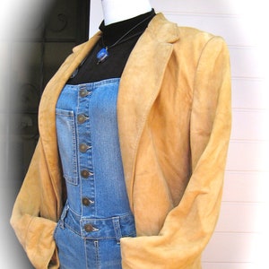 May include: A tan suede jacket with a blue denim overall jumpsuit underneath. The jacket has a long sleeve and a collar. The overall jumpsuit has a button closure and pockets.