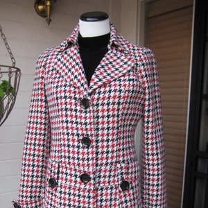 May include: A red, black, and white houndstooth patterned jacket with a black mock neck shirt underneath. The jacket has a button closure and two pockets.