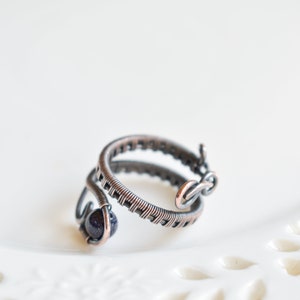 May include: A copper wire ring featuring a dark blue stone. The ring is crafted from two intertwined wires, one of which is wrapped around the stone.