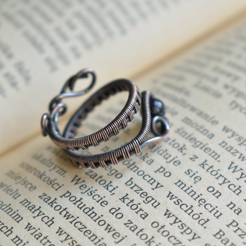 May include: A double-wrapped copper wire ring with a dark purple bead. The ring is on a book with text in a foreign language.