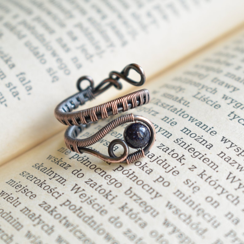 May include: A copper wire ring featuring a dark blue gemstone. The ring is wrapped in a woven pattern and has a heart-shaped design.