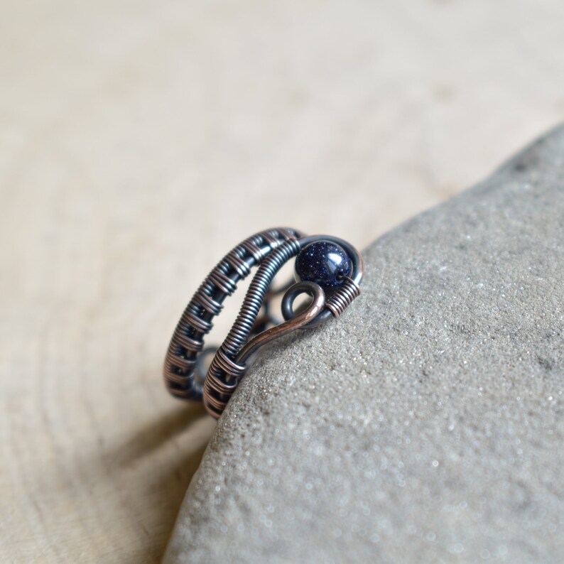 May include: A copper wire ring featuring a blue gemstone. The ring is crafted from multiple strands of wire that are interwoven. The gemstone is a round, blue stone with a speckled texture.