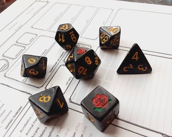 May include: A collection of black polyhedral dice with gold numbering and accents, including a triangular die and dice with a red rose emblem. The dice are arranged on a white game sheet with printed text.