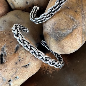 May include: Two silver cuff bracelets featuring a complex, woven chain-link design. The metal has a polished finish with darkened recessed areas to highlight the intricate braided pattern of the links. Each bracelet is an open-ended cuff style.