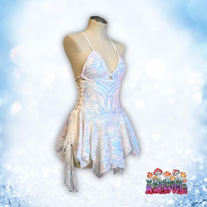 Puede incluir: Un vestido blanco de lentejuelas iridiscentes con un corsé de encaje y una falda con volantes. El vestido tiene una borla blanca que cuelga del lado. El vestido está en un maniquí blanco. El texto "MadebyVibe" está en la esquina inferior derecha de la imagen.