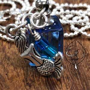 Peut inclure: Un collier en argent avec un pendentif en forme de flacon de parfum en verre bleu et un pendentif en argent en forme de poisson. Le collier est sur une surface en bois.