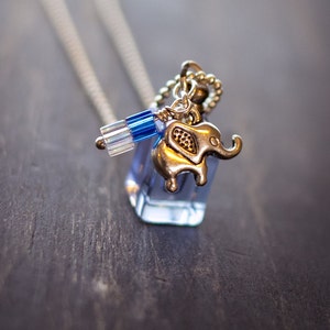 May include: A silver elephant charm hangs from a silver chain necklace. The charm is attached to a clear glass vial with blue and iridescent beads.