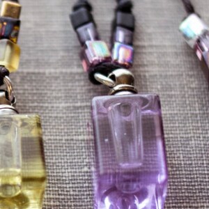 Puede incluir: Tres frascos de perfume de vidrio con tapas plateadas y cordones negros con cuentas de colores. Los frascos son transparentes y están llenos de líquido. Los frascos cuelgan de los cordones.