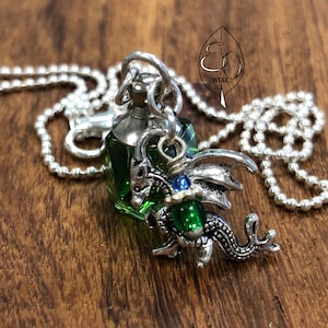Könnte beinhalten: Eine silberne Kettenkette mit einem grünen Glasflaschen-Anhänger und einem silbernen Drachen-Anhänger. Der Drachen-Anhänger hält einen grünen Edelstein.