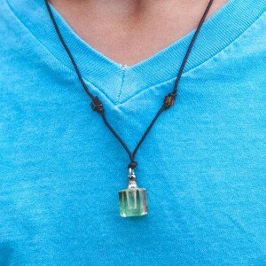 Op de afbeelding: Een groene glazen hanger aan een zwarte koordsnoer. De ketting wordt gedragen door iemand die een lichtblauw shirt draagt.