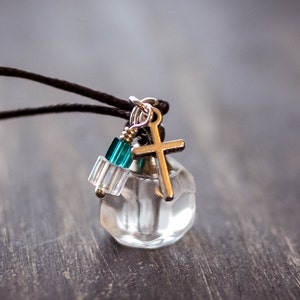 May include: A clear glass vial with a silver cross charm and teal and clear glass beads hanging from a black cord.