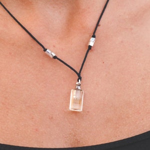 Puede incluir: Un collar de cordón negro con un pequeño colgante de vidrio transparente en forma de botella. La botella tiene una tapa plateada y está llena de un líquido ámbar claro. El collar tiene dos cuentas plateadas.