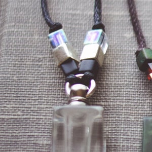 May include: A silver and black necklace with a clear glass bottle pendant. The bottle has a silver cap and a black cord.