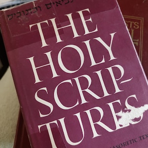 May include: A burgundy book titled "THE HOLY SCRIPTURES" with white lettering. The book has Hebrew text at the top and the text "ACCORDING TO THE MASORETIC TEXT" at the bottom. Another book is partially visible in the background.