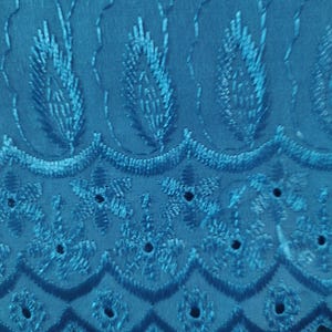 May include: Close-up of teal fabric with intricate embroidered floral and leaf patterns. The fabric features a scalloped edge with small circular cutouts, creating a delicate and textured design. The embroidery adds depth and visual interest to the textile.