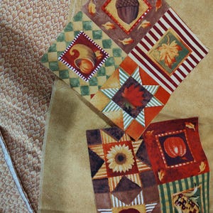 May include: A collection of fabric squares featuring autumn-themed prints including acorns, cornucopias, maple leaves, sunflowers, pumpkins, and apples. The patterns are set against backgrounds of green checkered, striped, and solid tan fabric textures.