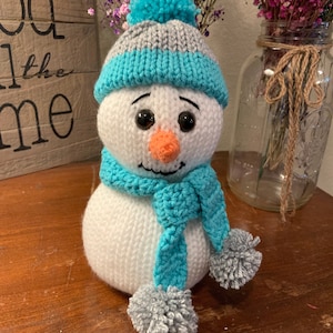 Puede incluir: Un muñeco de nieve de crochet blanco con un gorro de punto azul y gris, una bufanda azul con pompones grises y una nariz de zanahoria.