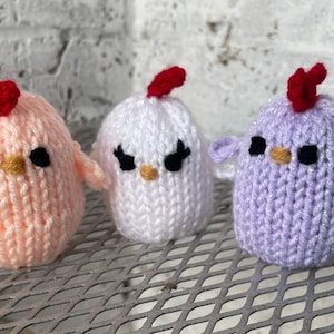 May include: Three knitted chick toys, one pink, one white, and one purple, with red combs and black eyes.