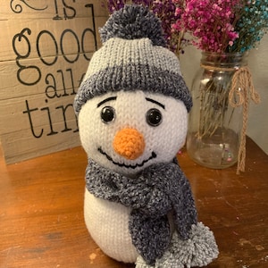 Puede incluir: Figura de muñeco de nieve hecha a mano con cuerpo blanco, que lleva un gorro y una bufanda de punto a rayas grises y blancas. El muñeco de nieve tiene una nariz de zanahoria, ojos negros y una sonrisa cosida. La bufanda tiene pompones grises.