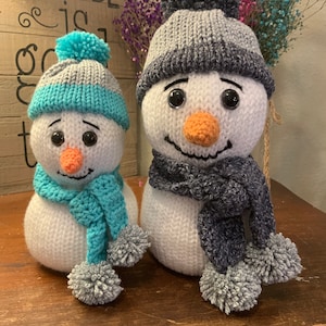 Puede incluir: Dos muñecos de nieve de crochet con sombreros grises y blancos, uno con una bufanda azul y el otro con una bufanda gris. Ambos muñecos de nieve tienen ojos negros y narices naranjas.