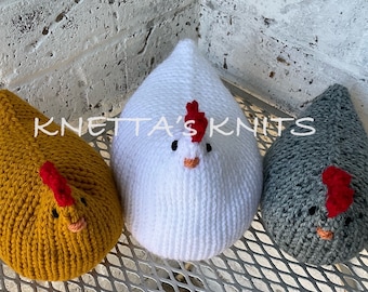 May include: Three knitted chicken plushies in different colours: yellow, white, and grey. Each plushie has a red comb and a small orange beak. The plushies are sitting on a metal surface with the text "KNETTA'S KNITS" visible in the background.