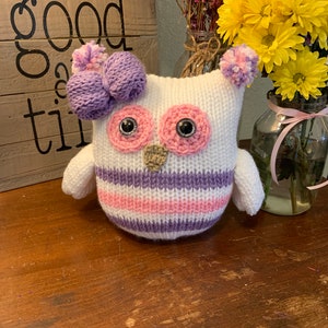 May include: A white knitted owl with purple and pink stripes, a purple bow, and pink eyes. The owl is sitting on a wooden surface.