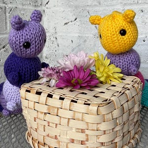 May include: Two crocheted caterpillar toys, one purple and one yellow, sit beside a woven basket filled with flowers. The caterpillars have large black eyes and are made of yarn. The basket is rectangular and has a natural wood color.
