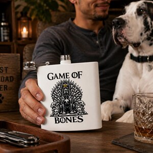 May include: A white flask with the words "GAME OF BONES" and a dog-themed design. The flask is held by a person, with a Great Dane dog in the background. A wooden sign that says "BEST DOG DAD EVER" is also visible.