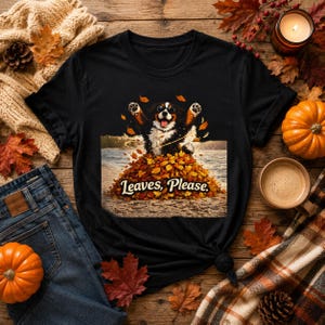 May include: A black short-sleeved t-shirt featuring a graphic of a Bernese Mountain Dog wearing sunglasses, jumping into a large pile of colorful autumn leaves. The text "Leaves, Please." is printed in white script below the dog illustration.