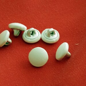 May include: Six white plastic buttons with a silver metal shank. The buttons are round and have a smooth surface.