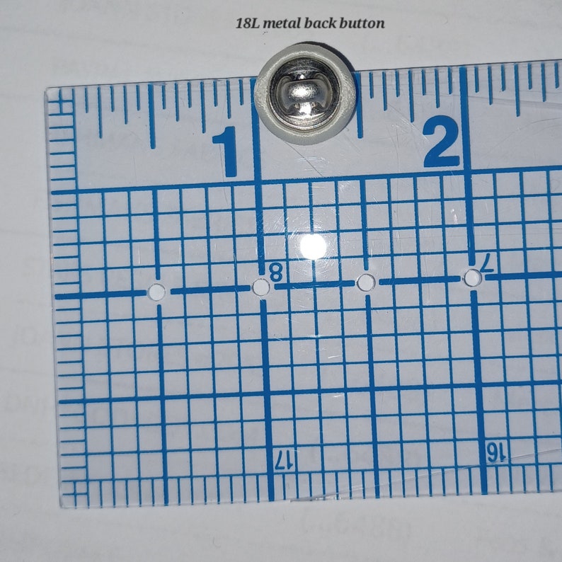 May include: A silver metal back button with a white plastic base, measuring approximately 1 inch in diameter. The button is shown on a ruler with a grid pattern.