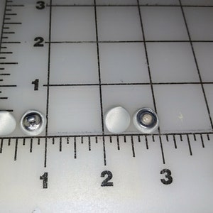 May include: Four white buttons, two with a smooth surface and two with a metal shank. The buttons are arranged in a row on a white surface with a ruler.