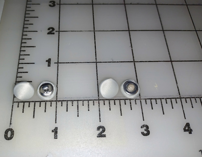 May include: Four white buttons, two with a smooth surface and two with a metal shank. The buttons are arranged in a row on a white surface with a ruler.
