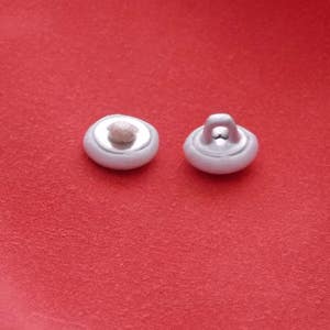 May include: Two white plastic buttons with a loop on one button. The buttons are on a red surface.