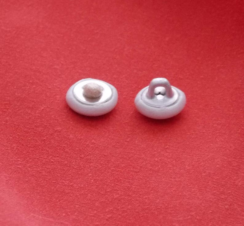 May include: Two white plastic buttons with a loop on one button. The buttons are on a red surface.