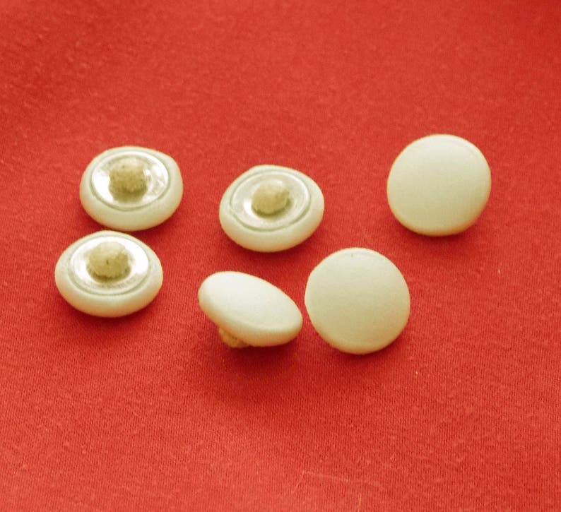 May include: Six white plastic buttons with a silver ring around the center. The buttons have a raised center and are arranged on a red surface.