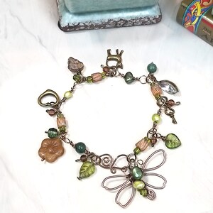 May include: A copper wire bracelet with charms, including a butterfly, leaves, a heart, a key, and beads in shades of green, brown, and peach.